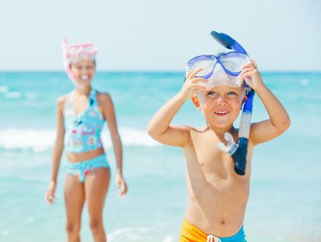 Happy young boy with snorkeling equipment on sandy tropical beach, his sister background.