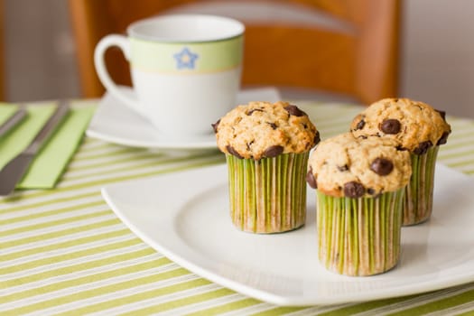 Three chocolate chip muffins on white plate and green striped tablecloth at breakfast