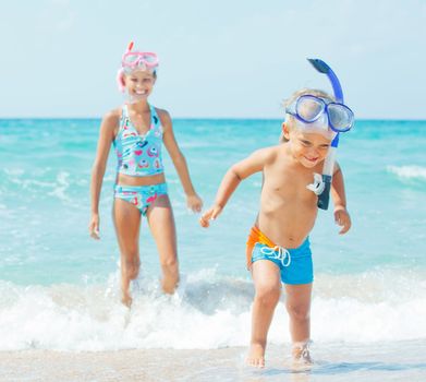 Happy young boy with snorkeling equipment on sandy tropical beach, his sister background.