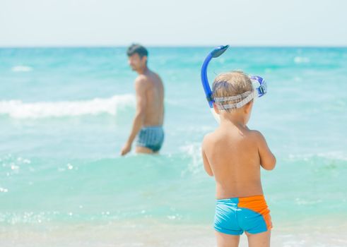 Cute little boy with snorkeling equipment on tropical beach, his father background.