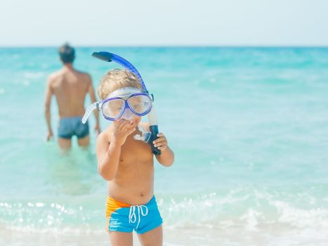 Cute little boy with snorkeling equipment on tropical beach, his father background.