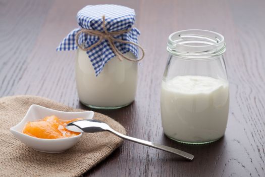 Homemade milk yogurt with peach or apricot jam in porcelain bowl and metal spoon served on linen napkin, wooden table