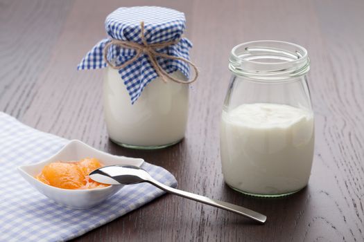 Homemade milk yogurt with peach or apricot jam in porcelain bowl and metal spoon served on linen napkin, wooden table