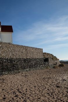 A high solid stone wall with cubed crates of stone reinforcements at sand level on a beach.