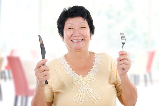Happy Asian senior mother ready for her Mother's day meal, holding fork and knife inside restaurant.