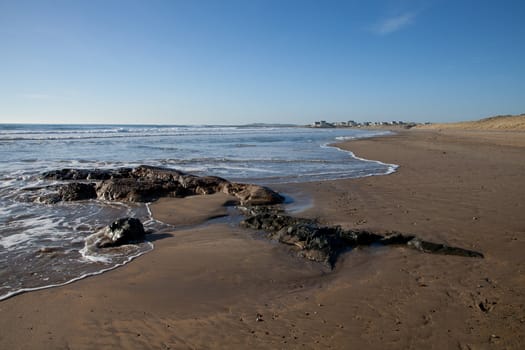 Rhosneigr beach with waves lapping on a rock and an empty beach with the village in the distance, Wales coast path, Anglesey, Wales, UK.