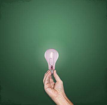 Hand holding a bright incandescent lightbulb in front of a green chalkboard with plenty of copy space