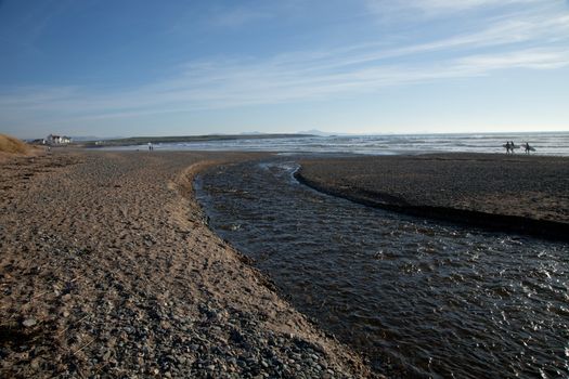 A river flows into the sea with surfers on the shore at Rhosneigr beach, Wales coast path, Anglesey, Wales, UK.