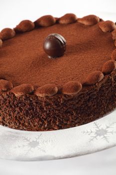 Photo of a delicious chocolate cake with truffle on top. Cake is on a plate with icing sugar with snowflake accents. Focus is on the front edge.
