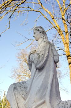 Dirty old white angel statue without wings. Trees without leaves in autumn.