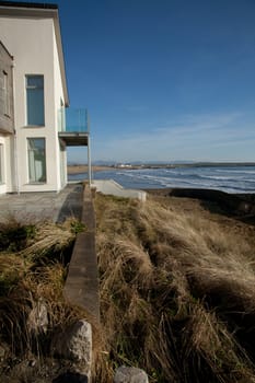 A wall leads to the exterior of a building with a first floor balcony overlooking the sea and beach in the distance.