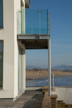 A first floor balcony with glass railings overlooks the sea and beach with surfers at the shore and dunes leading to mountains in the distance.