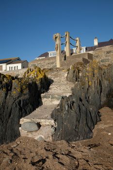 Concrete steps set into rocks on a beach leading to a wooden post and rope railings with house roofs in the distance.