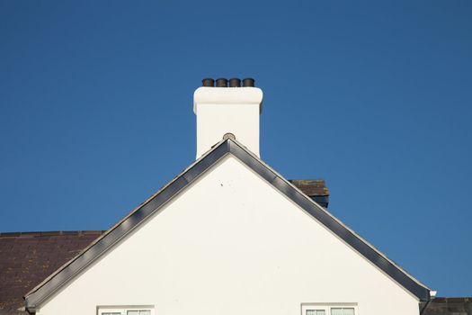 A white painted gable end with plastic barge boards a chimney and pots with a slate roof against a blue sky.