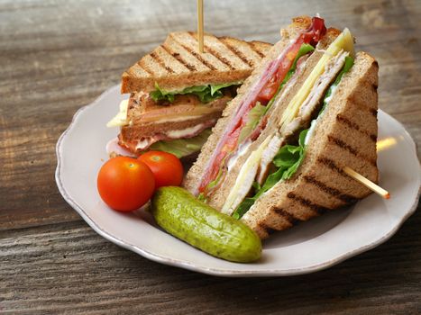Photo of a club sandwich made with turkey, bacon, ham, tomato, cheese, lettuce, and garnished with a pickle and two cherry tomatoes.