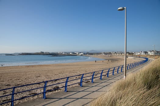 A footpath and blue railings stretch around the sandy beach at Trearddur bay, Anglesey, Wales, UK.