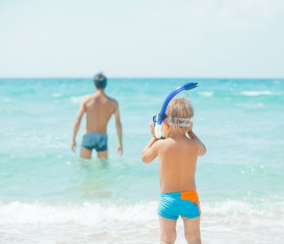 Cute little boy with snorkeling equipment on tropical beach, his father background.