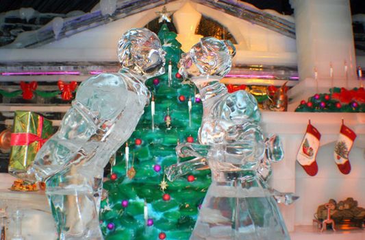 Made out of ice a couple kissing in front of a christmas tree.