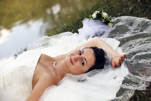 The beautiful bride with a wedding bouquet lays on a grass