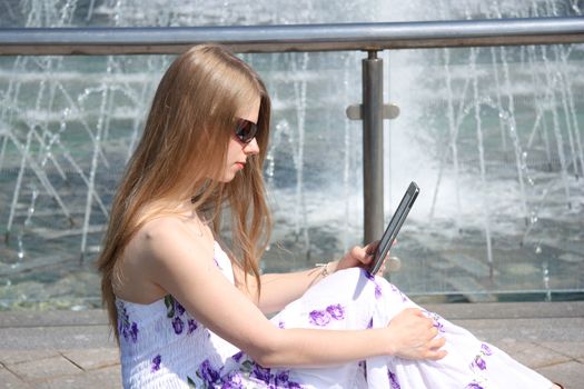 Young girl sitting on a bench and reading a digital book in a summer city