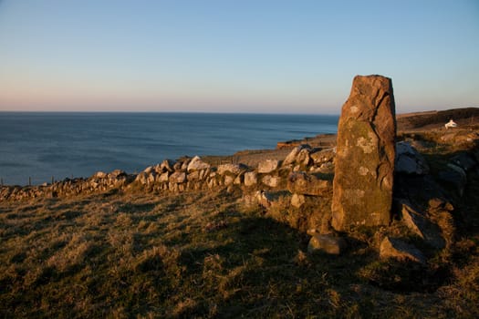 An ancient standing stone in the red evening light incorpoated into the corner of a stone wall with fields running down to the sea.