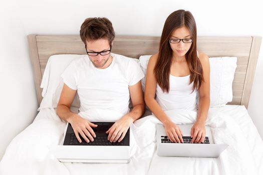 Serious young couple wearing glasses sitting side by side working on their laptops in bed. Beautiful young interracial couple, Asian woman, Caucasian man.
