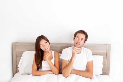 Thinking young couple sitting pensive in bed smiling and looking up at the ceiling for inspiration and ideas. Happy young interracial couple, Asian woman, Caucasian man.