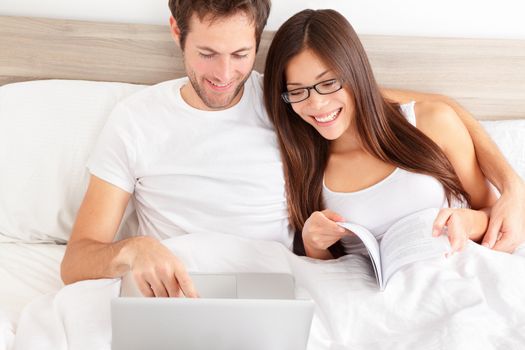 Happy affectionate young married couple sitting side by side in bed relaxing and laughing at information on the screen of their laptop. Happy young interracial couple, Asian woman, Caucasian man.