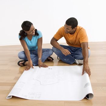 African American male and female couple sitting on floor looking at architectural blueprints.