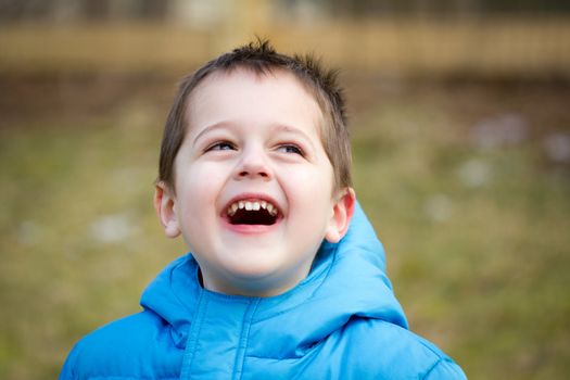 Portrait of a cute little brown-haired boy with a happy, joyful expression on his face.  He is wearing a blue coat and is playing outside.