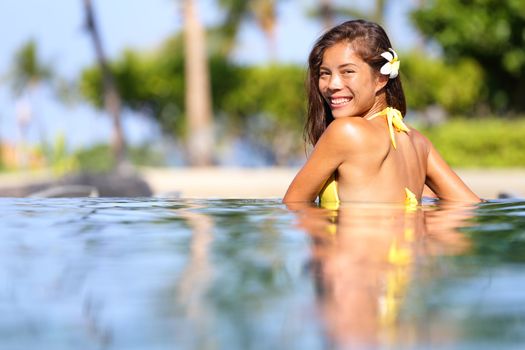 Vacation getaway woman swimming in a tropical pool. Smiling beautiful woman wearing a bikini relaxing in pool looking back over her shoulder across the inviting water at the camera. Multicultural girl