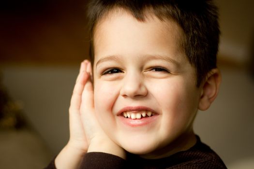 Close-up portrait of a cute little boy with brown hair and brown eyes smiling with his hands pressed to the side of his face.