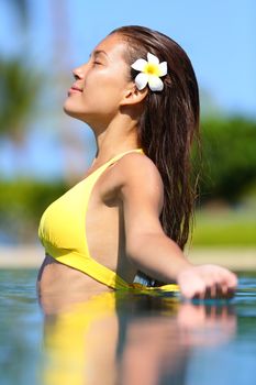 Spa woman in enjoyment meditating in water. Beautiful woman in bikini with frangipani flower in her hair standing meditating in a tropical pool with her arms outstretched and a serene expression.