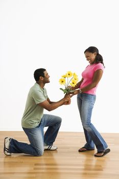 African American male giving pretty female a bouquet of yellow flowers while on bended knee.
