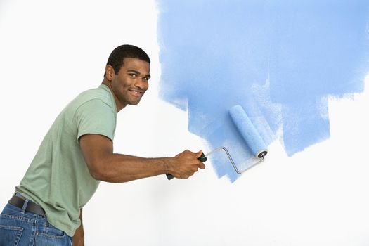 African American man painting wall blue smiling at viewer.