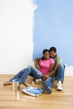 African American couple sitting together relaxing next to half-painted wall and painting supplies