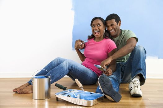African American couple sitting together relaxing next to half-painted wall and painting supplies