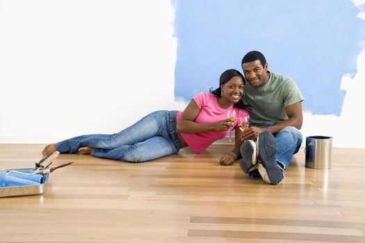African American couple relaxing together with wine next to half-painted wall and painting supplies.