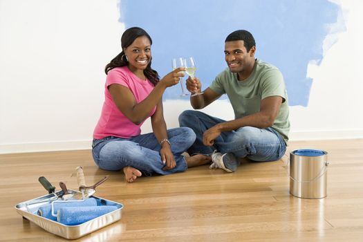 African American couple toasting with wine next to half-painted wall and painting supplies.