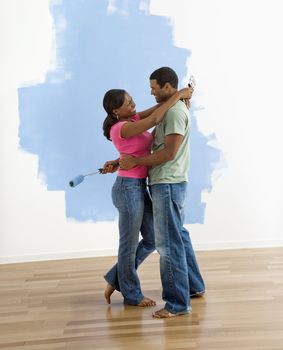 African American couple standing together hugging in front of half-painted wall.
