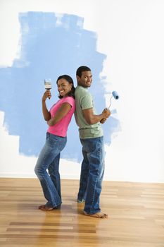 African American couple next to half-painted wall as they stand back to back with paint utensils.
