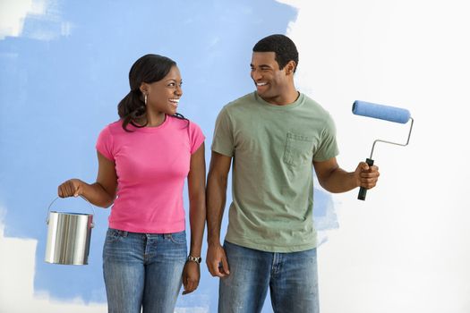 African American smiling couple next to half-painted wall with paint supplies.