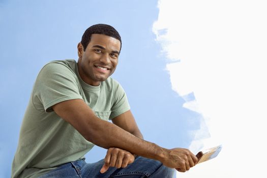African American young man sitting next to half-painted wall with paintbrush.