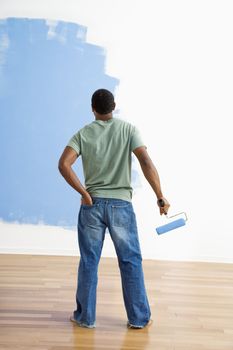 African American young man standing next to half-painted wall with paint roller.