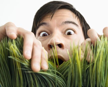 Asian young man looking through grass with fearful expression.