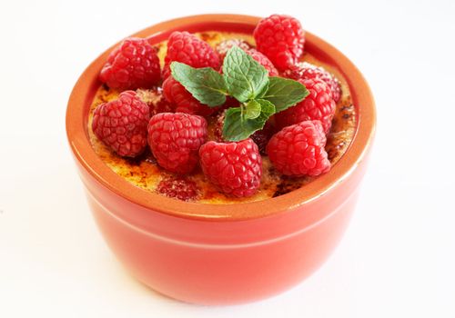 French creme brulee dessert with raspberries and mint covered with caramelized sugar in red terracotta ramekin on white background