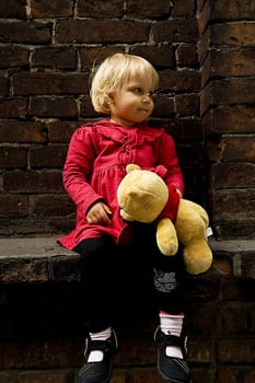 little blonde girl in red coat with teddy bear is seating on the window sill