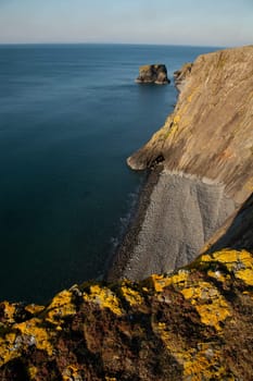 Yellow lichen covered rock on shale cliffs looking out towards a pebble beach and sea cliffs with the sea stack Trwyn y Tal in the distance. Wales coast path, Trefor, Lleyn peninsular, Wales, UK.