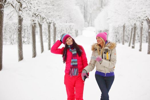 two women walk by winter alley snow trees on background