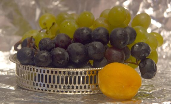 Apricot in the foreground in focus and two kinds of grapes in a silver bowl on a background